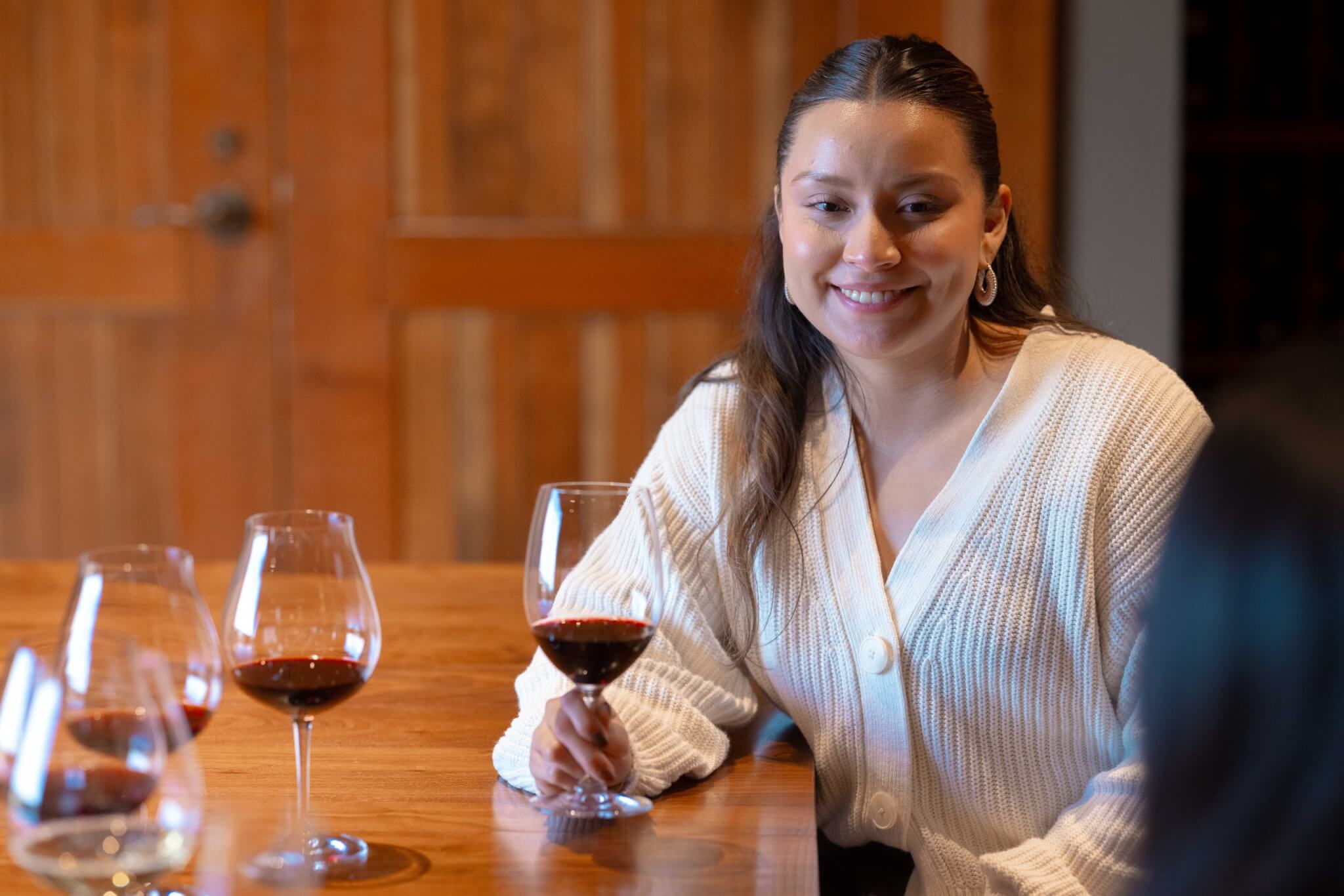 A woman in a white sweater smiles while holding a glass of red wine at a wooden table with several other wine glasses in Central Oregon.
