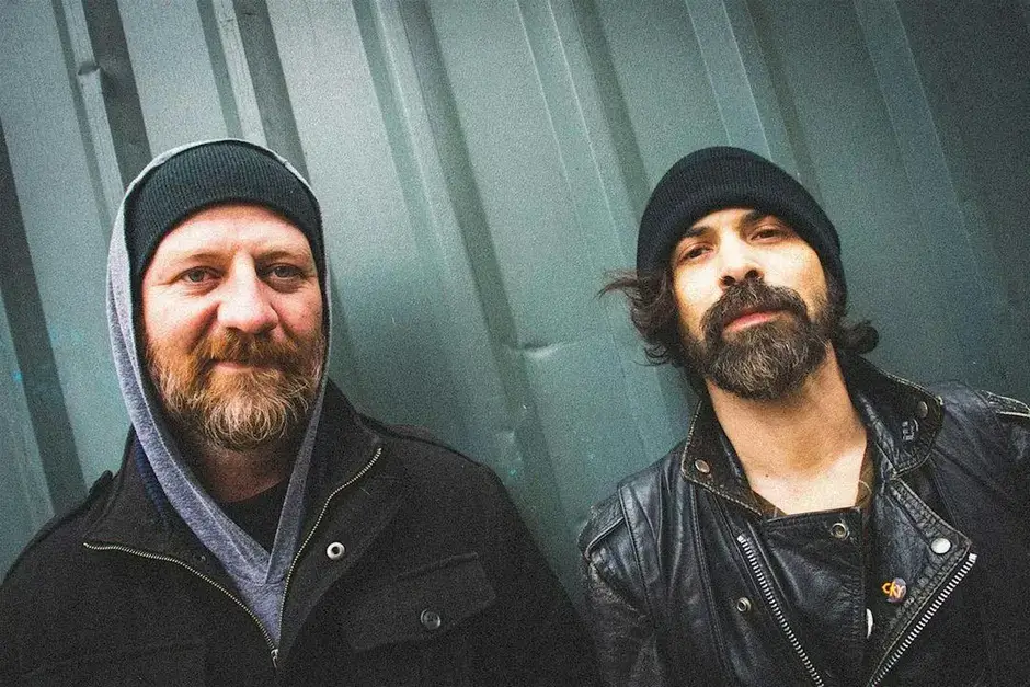 Two bearded men in black jackets and beanies stand against a corrugated metal wall in Central Oregon.