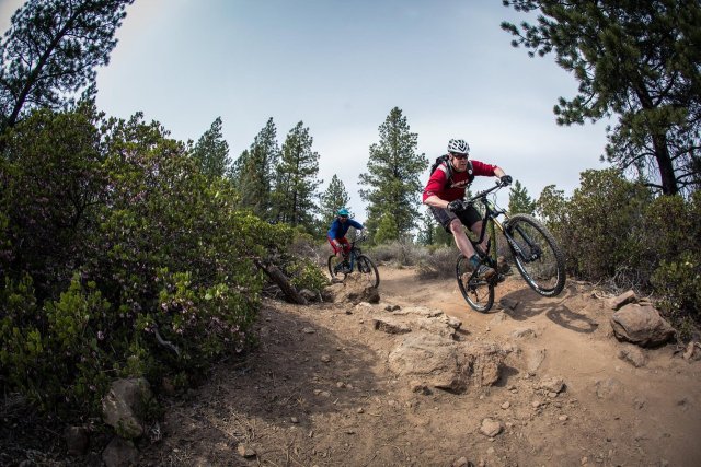 Two mountain bikers navigate a rocky trail through a lush forest, one in red and the other in blue, both wearing helmets and sunglasses in Central Oregon.