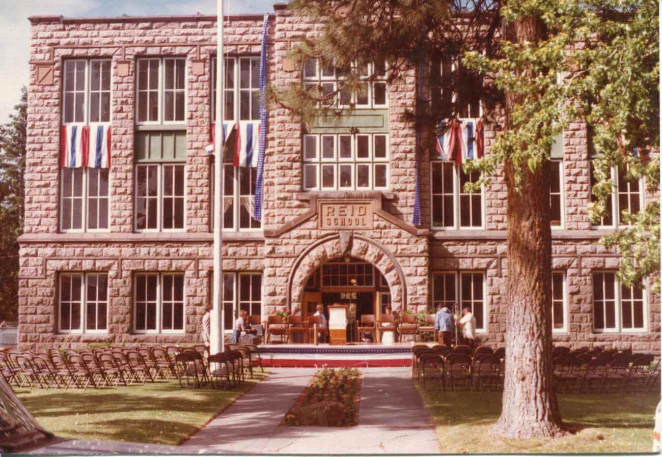 A historic stone building with a grand entrance, surrounded by neatly arranged rows of chairs and decorated with flags, suggesting a formal event or ceremony in Central Oregon.