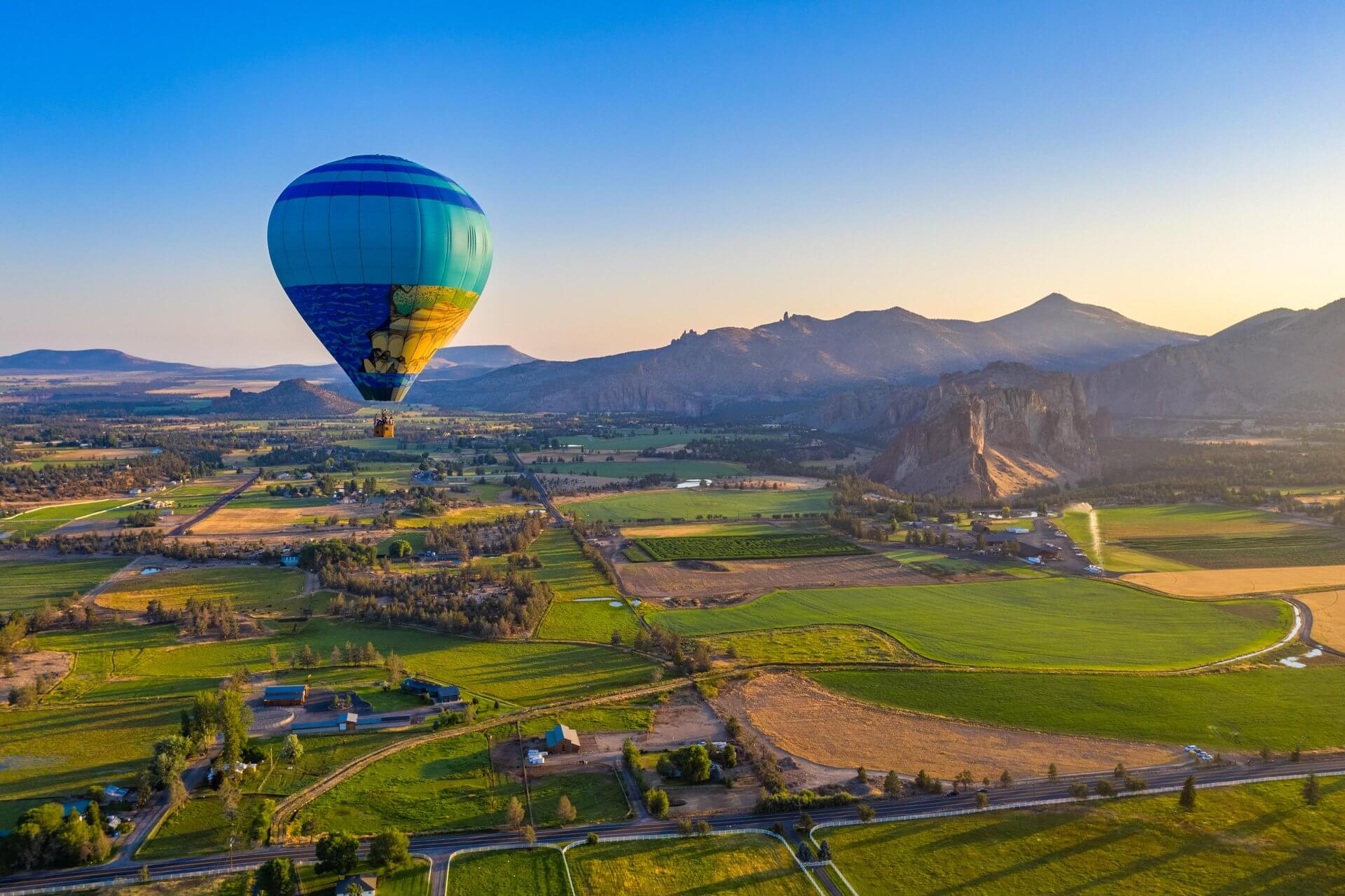 A vibrant hot air balloon soars over a lush green valley with rolling hills and distant mountains bathed in warm sunlight in Central Oregon.
