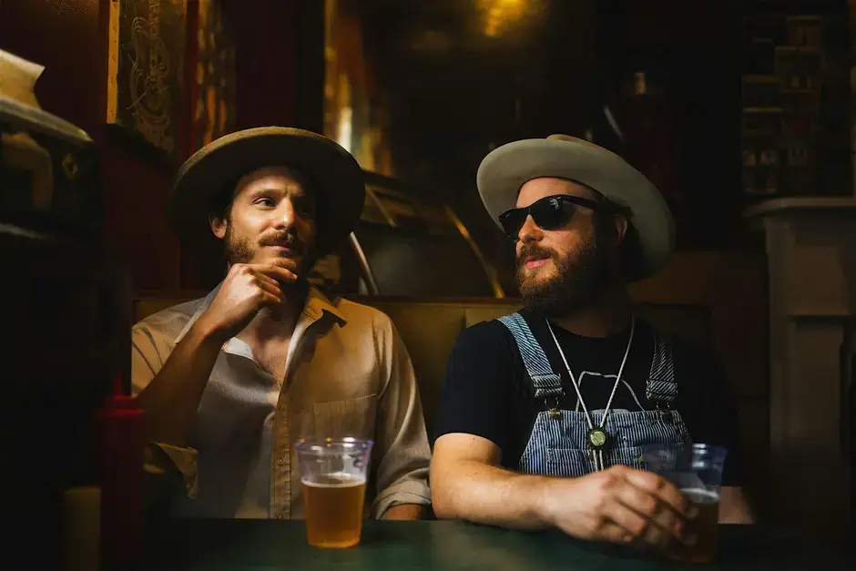 Two men in cowboy hats and sunglasses sit at a dimly lit bar, each holding a glass of beer in Central Oregon.