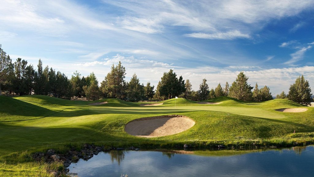 A serene golf course with undulating green fairways, sand traps, and a tranquil pond surrounded by lush trees under a clear blue sky in Central Oregon.