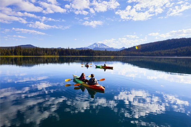 Kayakers paddle on a serene lake surrounded by mountains and forests under a clear blue sky in Central Oregon.