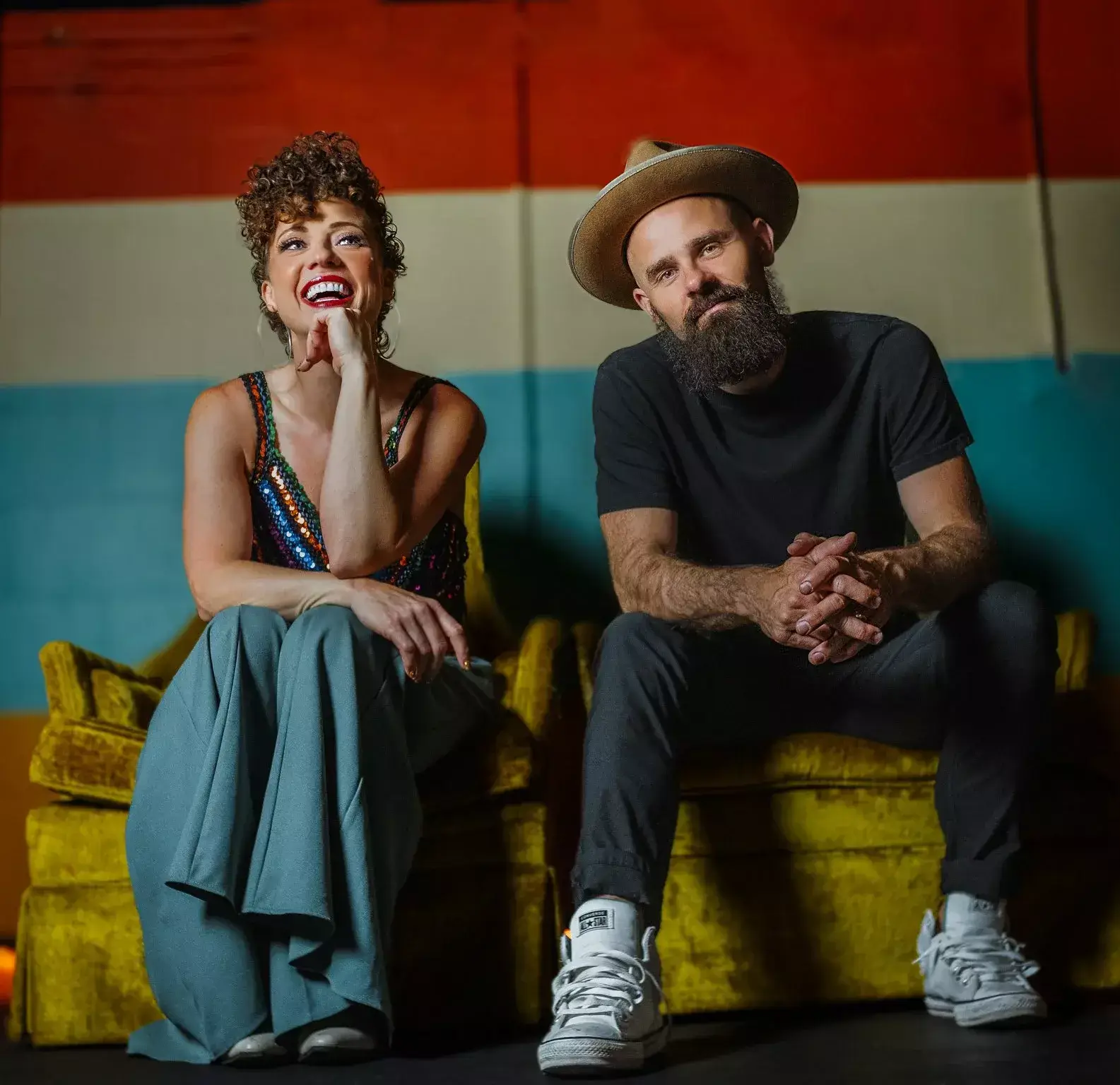A man and woman sit on a yellow velvet couch, laughing and looking at each other against a colorful striped backdrop in Central Oregon.