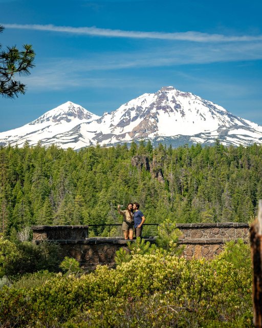 A couple takes a selfie on a stone wall overlooking a lush green forest with majestic snow-capped mountains in the distance in Central Oregon.