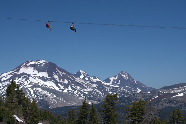 Two people ziplining across a scenic mountain valley with snow-capped peaks in the background in Central Oregon.