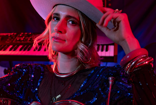 A woman wearing a wide-brimmed hat and a sequined dress, standing in front of a keyboard bathed in pink light in Central Oregon.