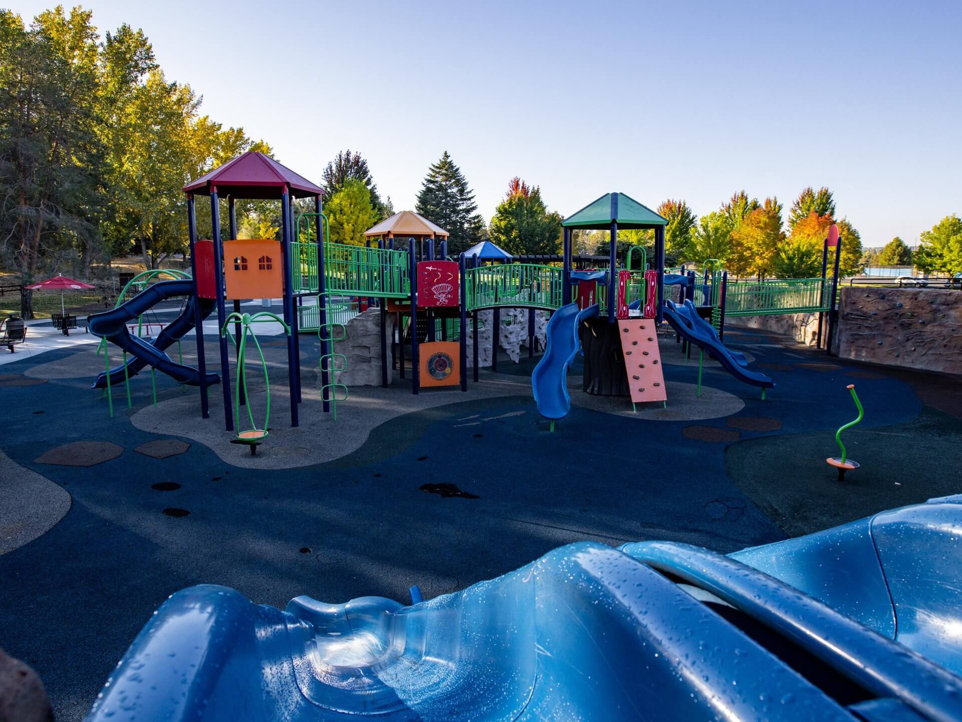 A vibrant playground with colorful slides, climbing structures, and playhouses surrounded by lush trees and a clear blue sky in Central Oregon.