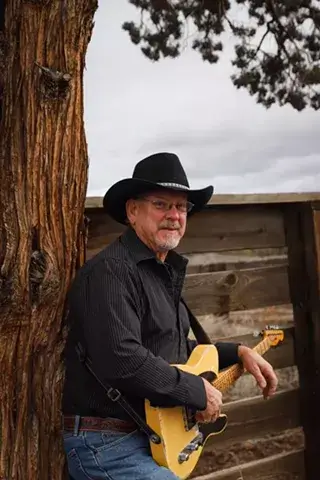 An elderly man wearing a cowboy hat and black shirt plays a yellow electric guitar against a backdrop of a weathered wooden fence and a large tree trunk in Central Oregon.
