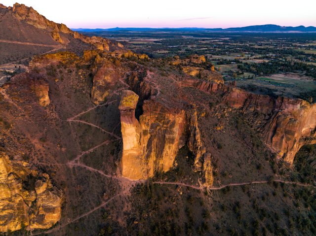 A rugged, golden-hued rock formation rises dramatically from a desert landscape at sunset, with winding trails leading up its slopes in Central Oregon.