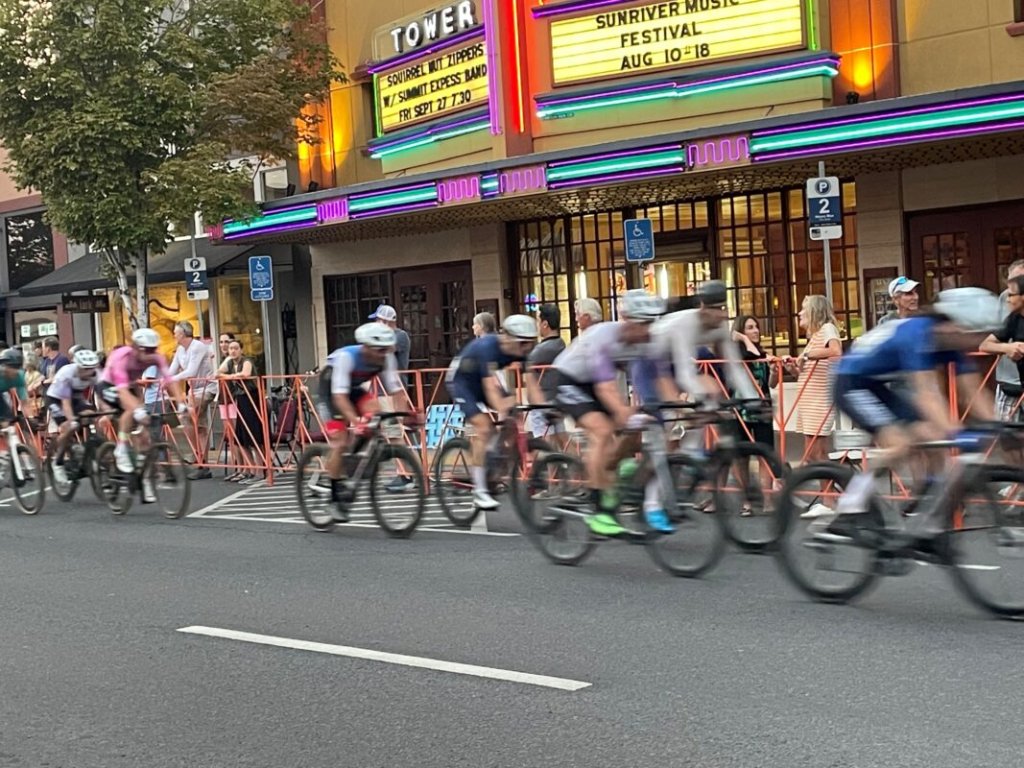 A group of cyclists in colorful jerseys races down a city street, passing by a vintage movie theater with neon signs advertising upcoming shows in Central Oregon.