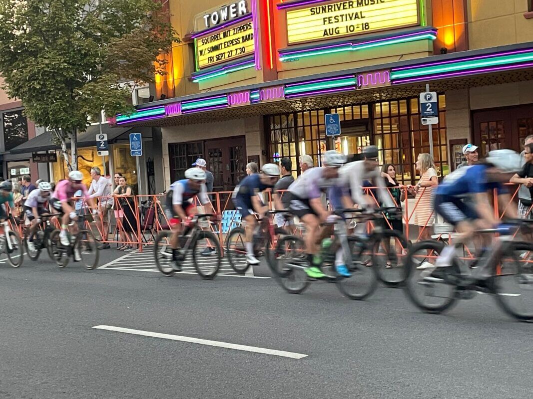 A group of cyclists in colorful jerseys races down a city street, passing by a vintage movie theater with neon signs advertising upcoming shows in Central Oregon.