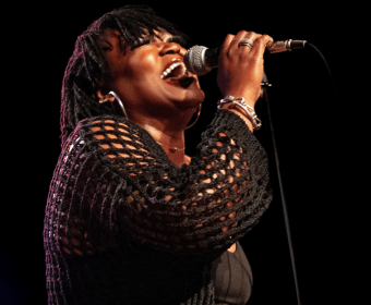 A passionate singer with dreadlocks performs on stage, wearing a black mesh top and holding a microphone close to her mouth in Central Oregon.