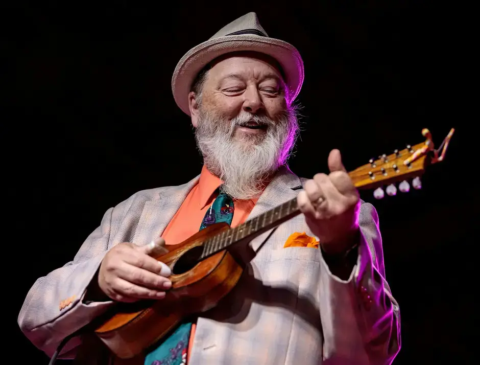 A bearded man in a suit and hat plays a small guitar with a joyful expression in Central Oregon.