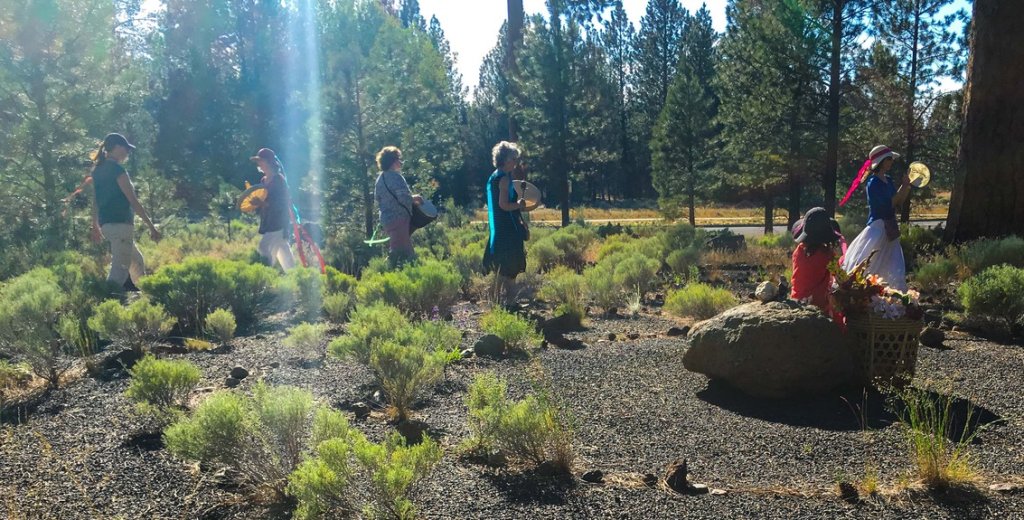 A group of people dressed in colorful costumes walk through a sunlit forest clearing, carrying baskets and musical instruments in Central Oregon.