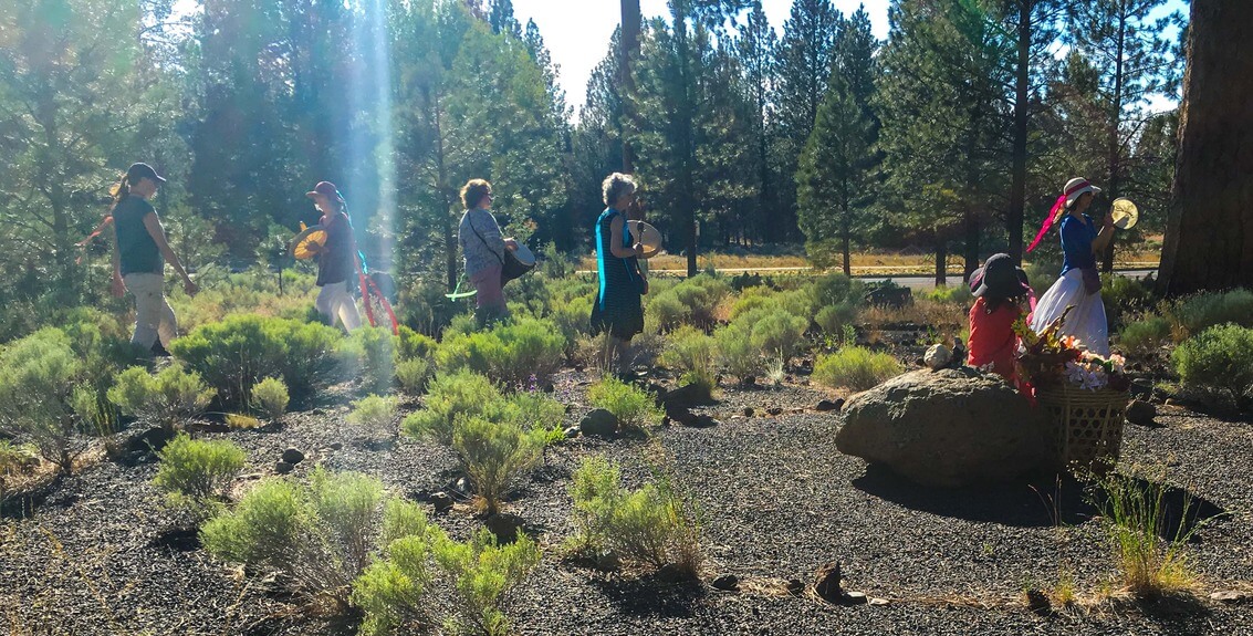 A group of people dressed in colorful costumes walk through a sunlit forest clearing, carrying baskets and musical instruments in Central Oregon.