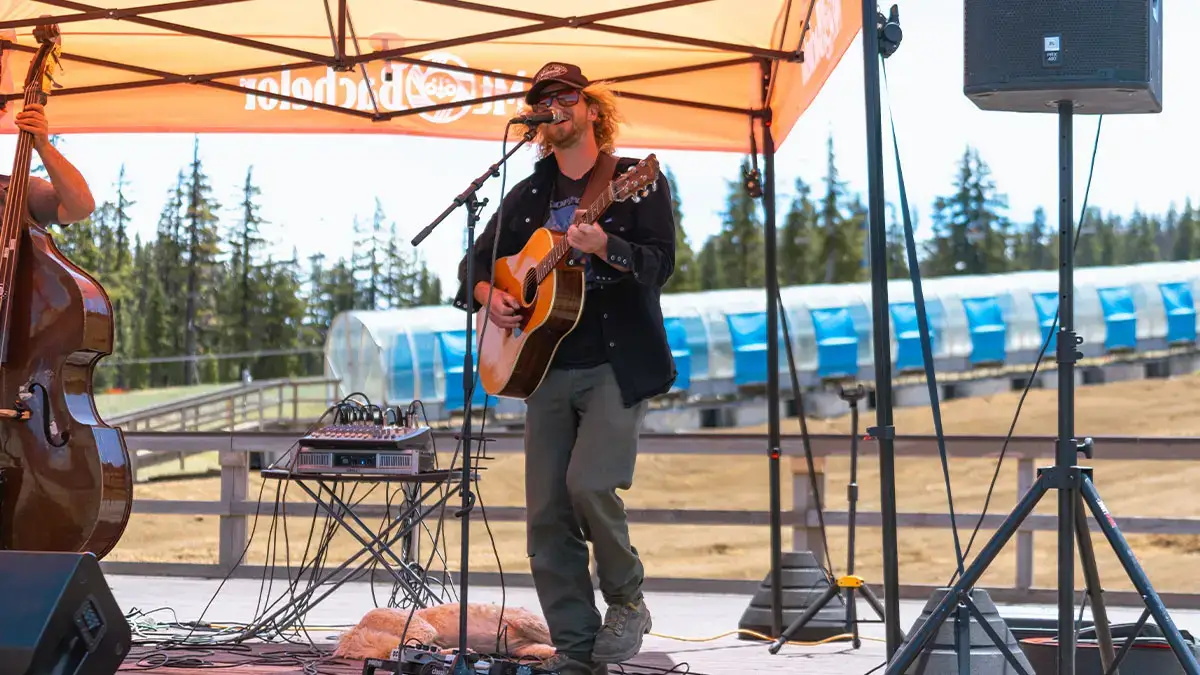 A musician with long hair and a beard plays an acoustic guitar on an outdoor stage under a bright orange tent, with a backdrop of lush green trees and a blue train carriage in Central Oregon.