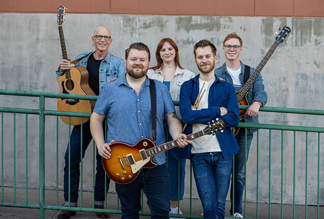 A group of five musicians, three men and two women, pose together with their guitars against a weathered concrete wall in Central Oregon.