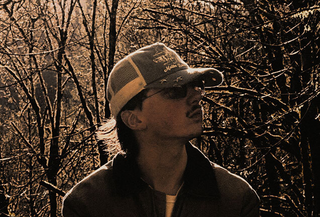 A young man wearing a cap and sunglasses stands in a forest during golden hour in Central Oregon.