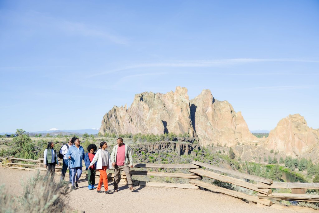 A diverse group of people enjoys a sunny day hiking along a scenic trail with towering rock formations in the background in Central Oregon.
