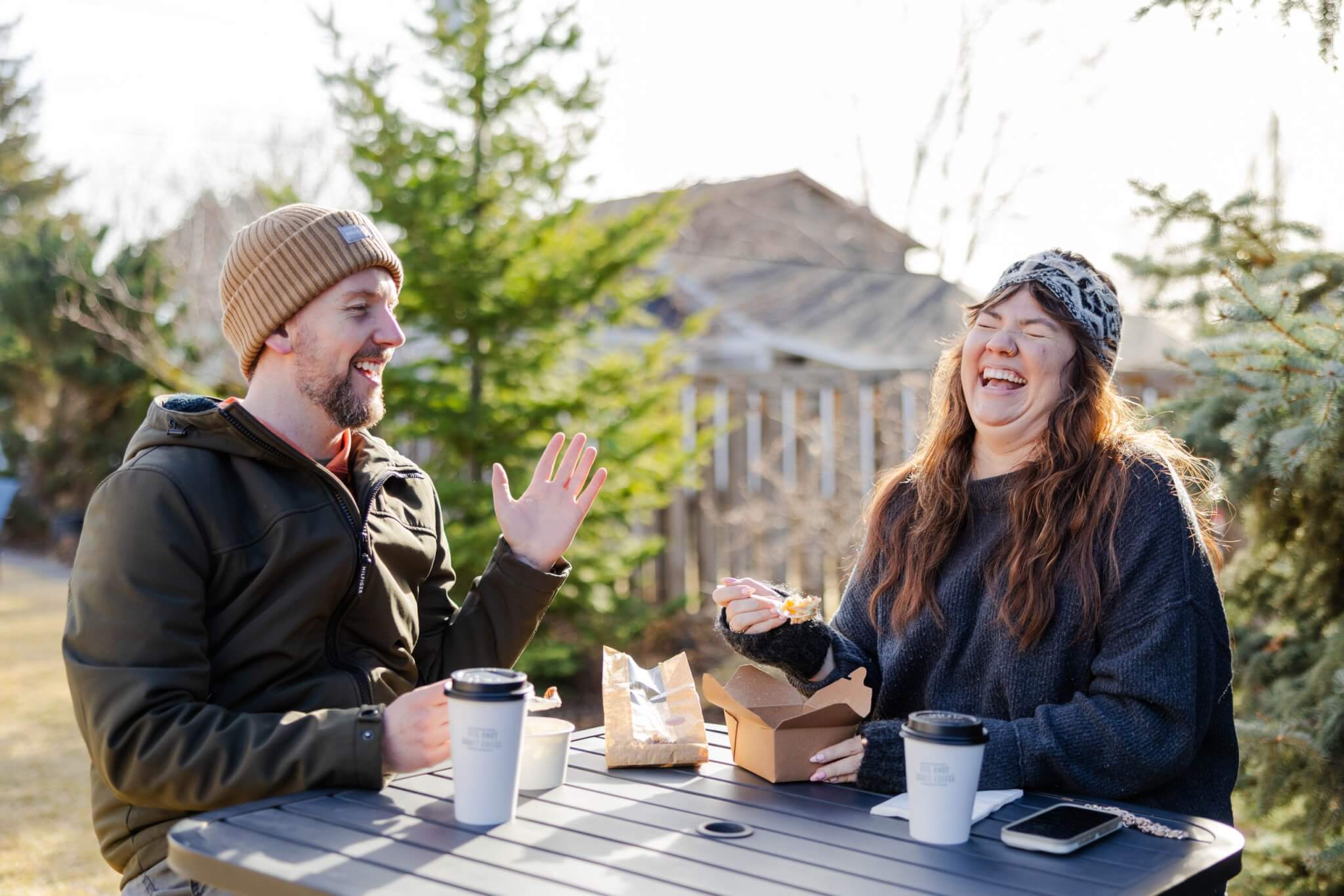 A couple enjoys a casual outdoor meal, laughing and chatting over coffee and takeout food at a picnic table surrounded by trees in Central Oregon.