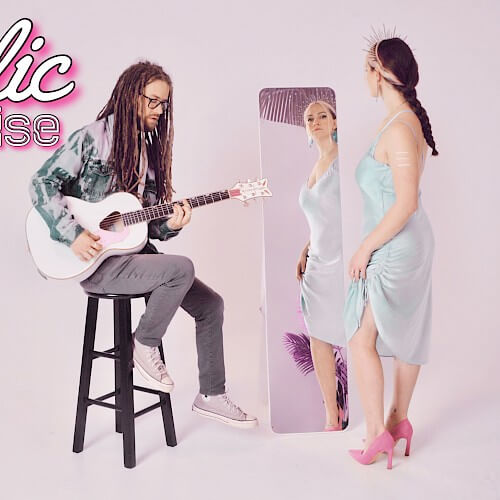 A man with dreadlocks plays guitar while a woman in a white dress stands in front of a mirror in Central Oregon.