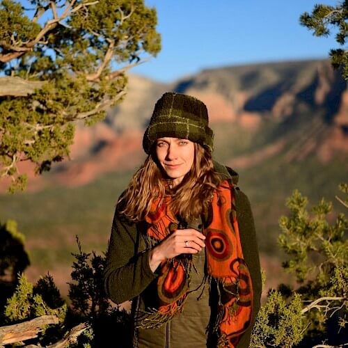 A woman wearing a green jacket, orange scarf, and hat stands amidst green trees with red rock formations in the background in Central Oregon.