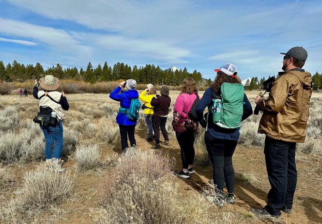 A group of hikers in colorful outdoor gear stands in a dry, grassy field, observing the landscape in Central Oregon.