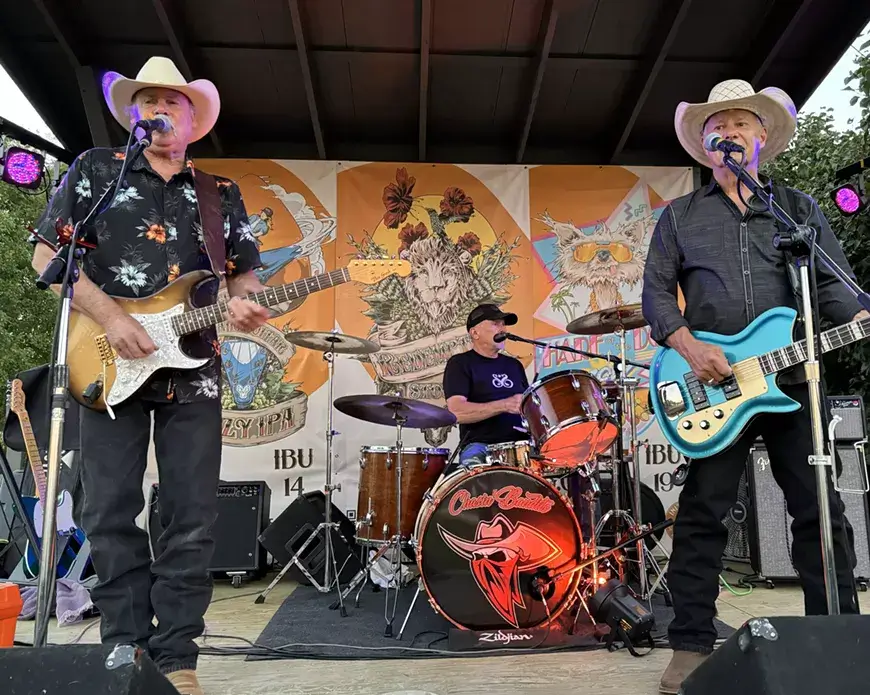 Two older men in cowboy hats and Hawaiian shirts play electric guitars on stage, accompanied by a drummer wearing a black t-shirt and a drum set with a red logo in Central Oregon.