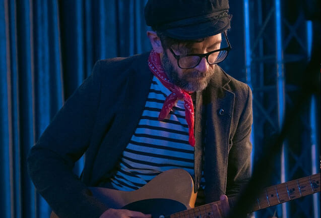 A musician wearing a striped shirt, cap, and glasses plays an electric guitar on stage in Central Oregon.