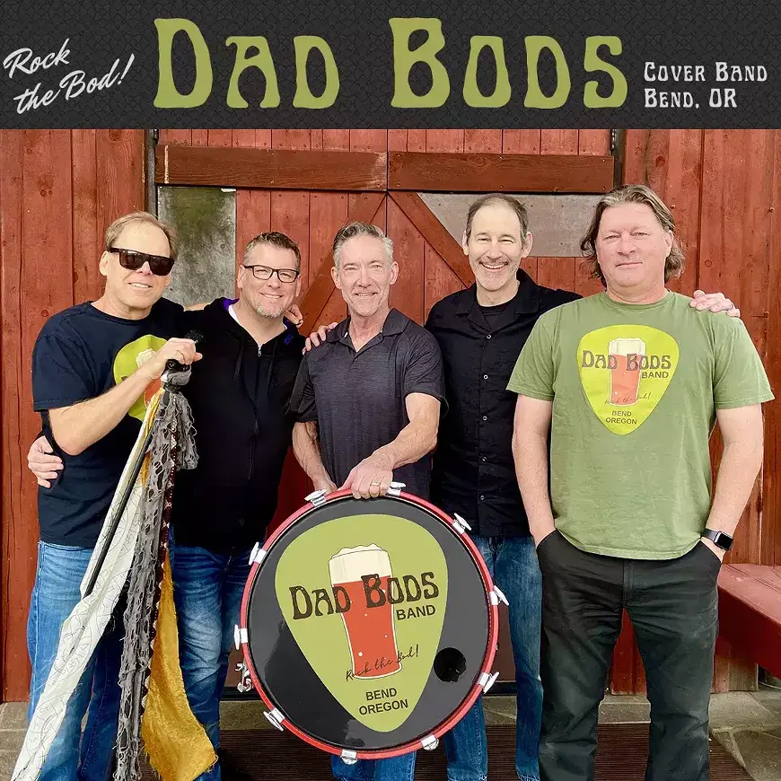 Five smiling men pose together with a drum featuring the Dad Bods logo, standing in front of a rustic wooden barn in Bend, Oregon in Central Oregon.