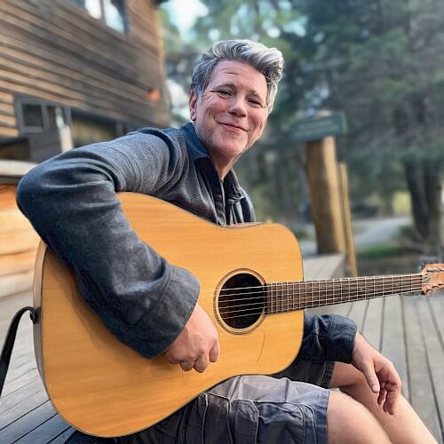 A man with gray hair smiles while playing an acoustic guitar on a wooden deck surrounded by trees in Central Oregon.