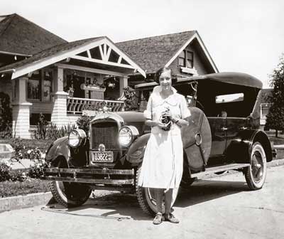 A woman in a white dress stands beside a vintage convertible car parked in front of a charming Craftsman-style house with a porch in Central Oregon.