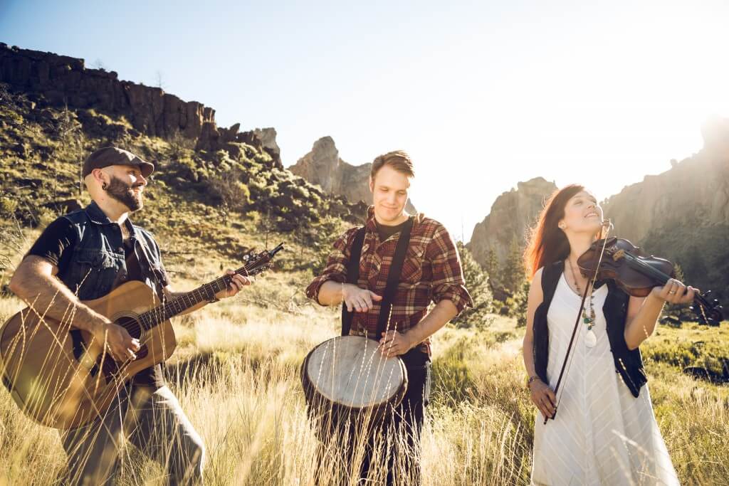 Three musicians play instruments in a golden field with rocky mountains in the background in Central Oregon.