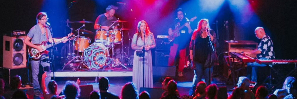 A vibrant live music performance featuring a band with guitars, drums, and keyboards, illuminated by colorful stage lights, as an enthusiastic audience watches from below in Central Oregon.