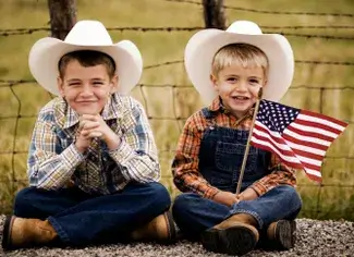 Two young boys dressed in cowboy attire, complete with white cowboy hats and plaid shirts, sit cross-legged on a gravel path, smiling broadly as they hold a small American flag between them in Central Oregon.