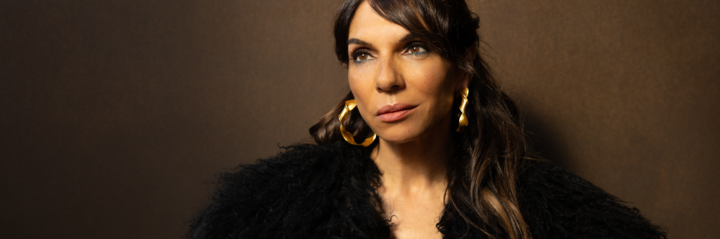 A woman with long dark hair wearing large gold hoop earrings and a black fur coat against a brown background in Central Oregon.