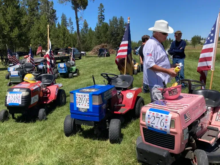 A row of lawnmowers decorated with American flags and patriotic designs are lined up on a grassy field, with people standing nearby in Central Oregon.