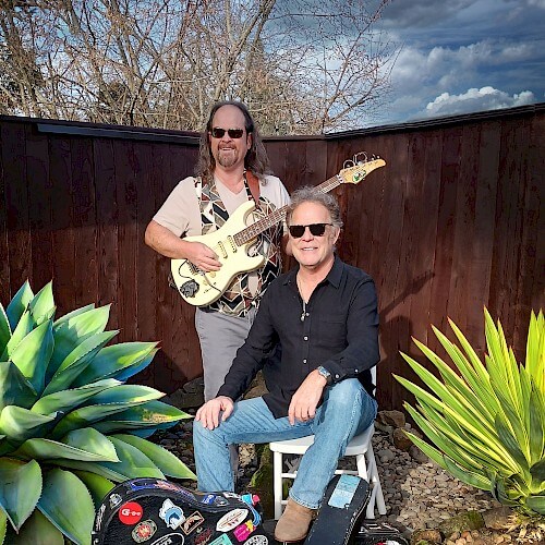 Two musicians, one playing guitar and the other seated with a guitar case, pose outdoors amidst lush greenery in Central Oregon.