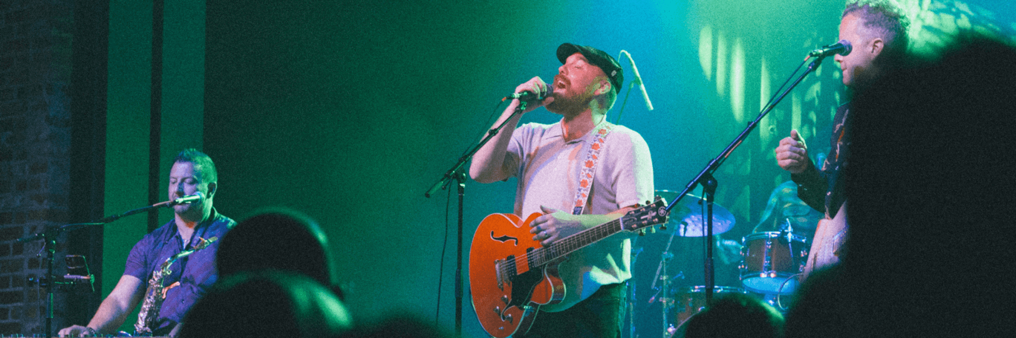 A lively band performs on stage with vibrant green lighting, featuring a lead singer with a guitar, a saxophonist, and a drummer in Central Oregon.