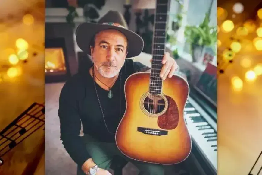 A musician wearing a hat and necklace sits by a fireplace with an acoustic guitar and a piano in the background in Central Oregon.