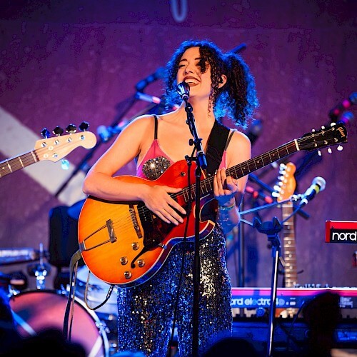 A musician with curly hair performs on stage, playing an electric guitar and singing into a microphone in Central Oregon.