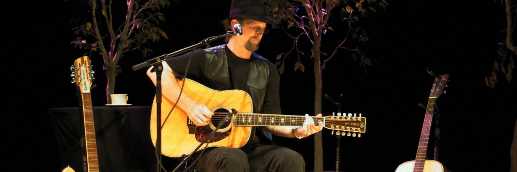 A musician wearing a black hat and vest performs on stage with an acoustic guitar, accompanied by two other guitars and a cup of coffee on a stand in Central Oregon.