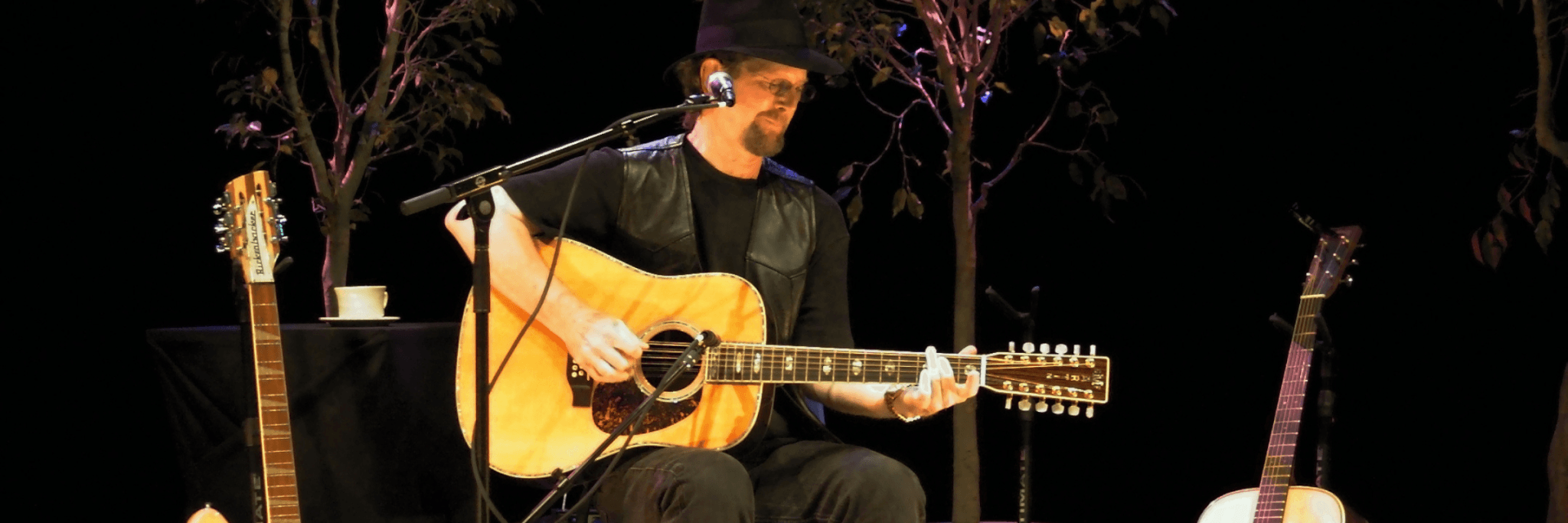A musician wearing a black hat and vest performs on stage with an acoustic guitar, accompanied by two other guitars and a cup of coffee on a stand in Central Oregon.