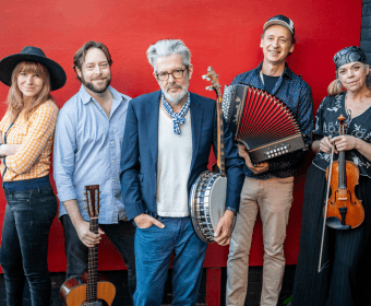 A group of five musicians, dressed in casual attire, stand together against a vibrant red wall, each holding a different musical instrument, including a guitar, banjo, accordion, violin, and fiddle in Central Oregon.