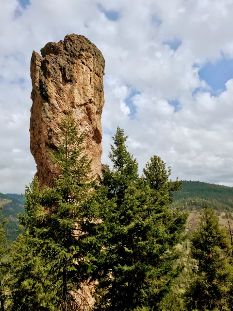 A towering rock formation rises majestically from a lush forest of evergreen trees, with a backdrop of rolling hills and a partly cloudy sky in Central Oregon.