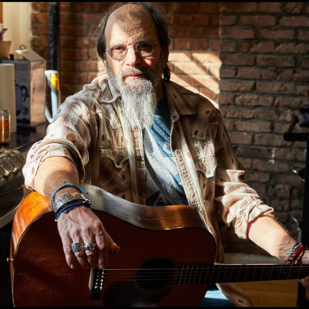 An elderly man with a long beard and glasses plays an acoustic guitar in a cozy room with a brick wall in Central Oregon.