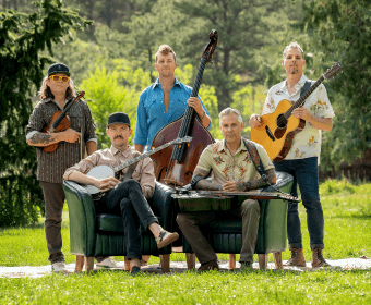 A group of five musicians, dressed in casual attire, pose together outdoors with their instruments, including a banjo, guitar, fiddle, upright bass, and resonator guitar in Central Oregon.