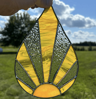 A vibrant yellow stained glass suncatcher with a teardrop shape and intricate geometric patterns, held against a backdrop of a clear blue sky and lush green fields in Central Oregon.